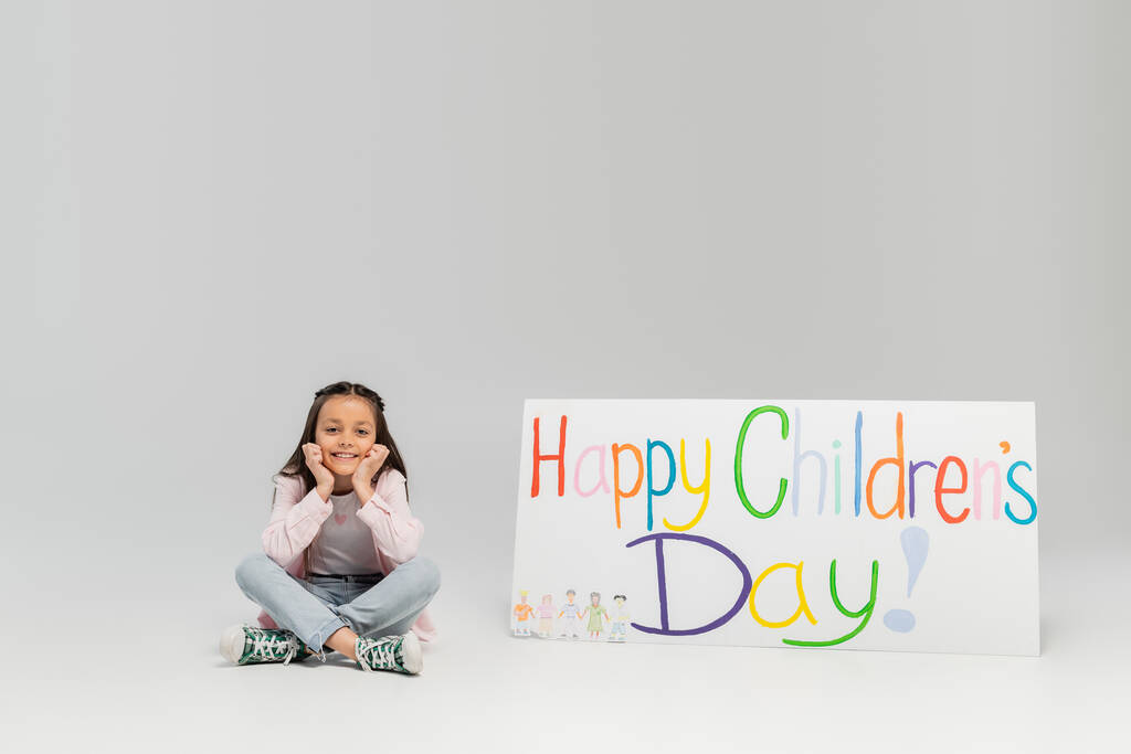 幸せな子供の日のレタリングとプラカードの近くのカメラを見て座っているカジュアルな服で喜びのプリティーガール6月のお祝いの間にグレーの背景にコピースペース - 写真・画像