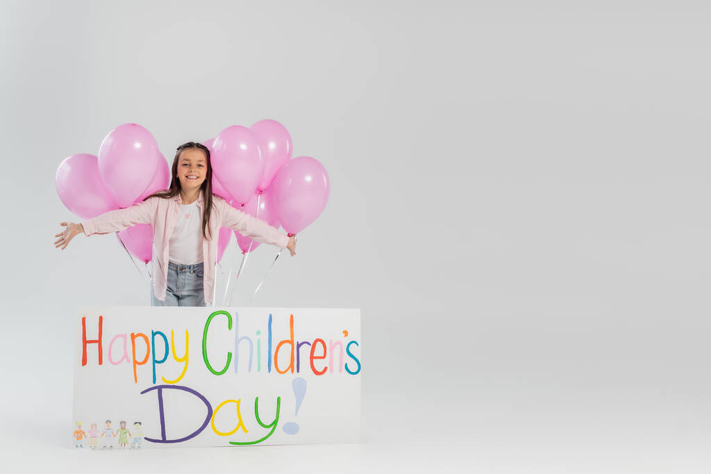 Χαρούμενο και preteen κορίτσι σε casual ρούχα κοιτάζοντας κάμερα κοντά σε ροζ εορταστικά μπαλόνια και πλακάτ με χαρούμενα γράμματα ημέρα των παιδιών, ενώ γιορτάζει σε γκρι φόντο - Φωτογραφία, εικόνα