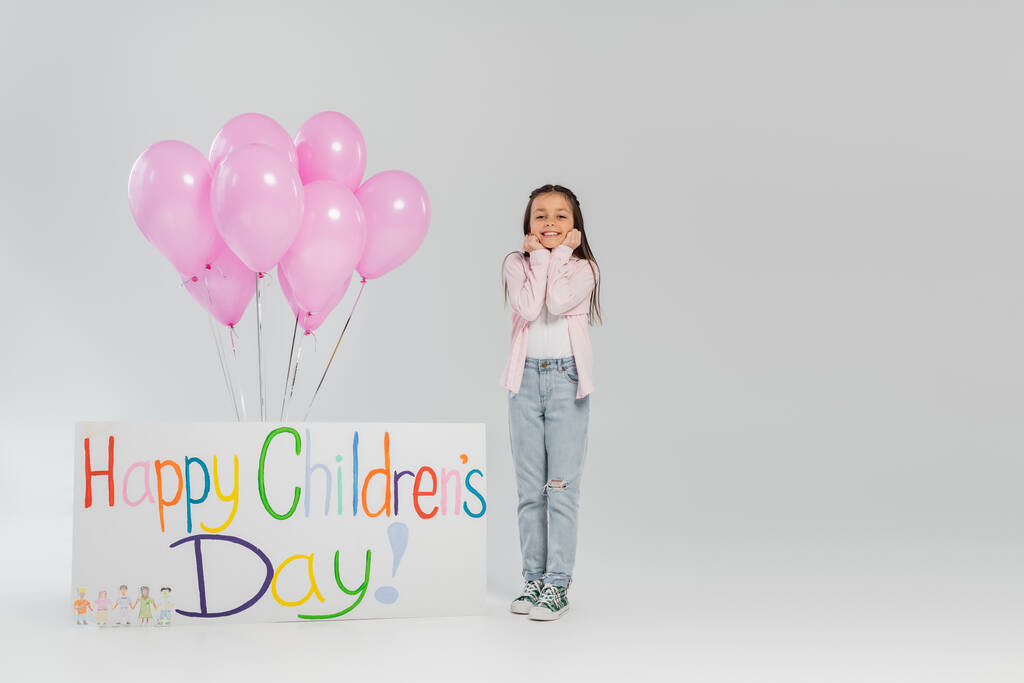 Pełna długość beztroski nastolatek dziewczyna w ubraniu casual patrząc w aparat, stojąc w pobliżu różowe balony i tabliczki z szczęśliwy dzień dzieci litery na szarym tle - Zdjęcie, obraz