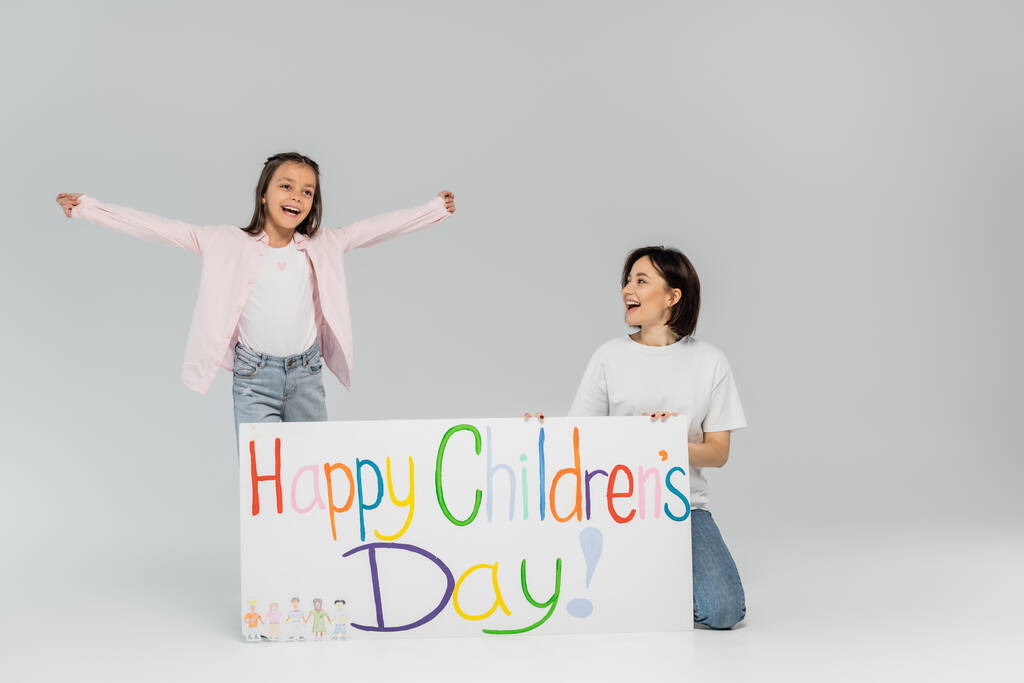 Excited preteen girl in casual clothes looking away while standing near mother and placard with happy children's day lettering during celebration on grey background - Photo, Image