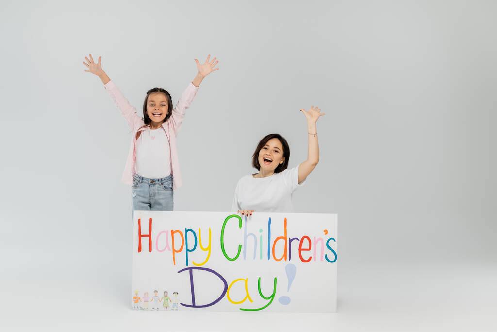 Aufgeregtes Mädchen in legerer Kleidung winkt in die Kamera neben Mutter und Plakat mit fröhlichem Kindertagsaufdruck bei Feier im Juni auf grauem Hintergrund - Foto, Bild
