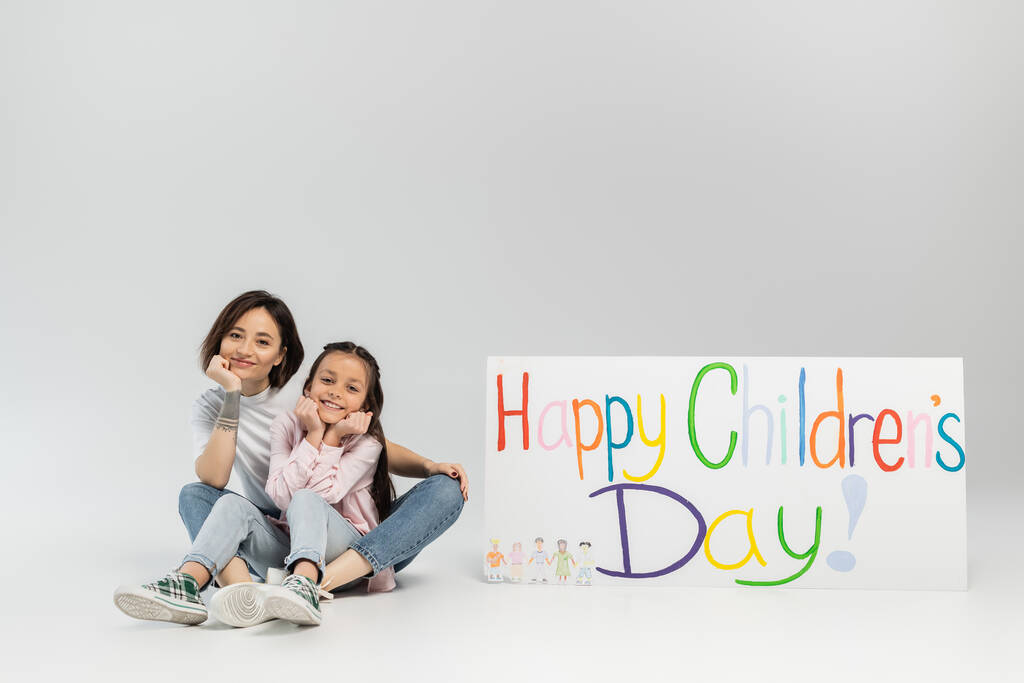 Full length of smiling tattooed mother looking at camera while sitting near daughter and placard with happy children's day lettering during celebration on grey background - Photo, Image