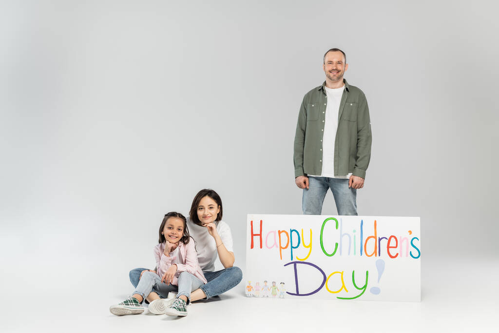 Smiling man looking at camera while standing near wife, preteen daughter and placard with happy children's day lettering during celebration on grey background - Photo, Image