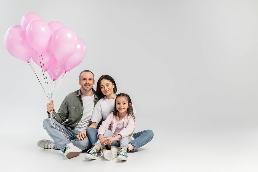 Usmívající se rodiče v ležérní oblečení objímání před dospívající dcera a držení slavnostní růžové balónky při oslavě mezinárodní děti den na šedém pozadí - Fotografie, Obrázek
