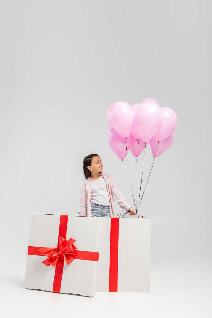 Niño preadolescente sonriente con ropa casual mirando globos rosados mientras está de pie en una gran caja de regalo durante la celebración del día de los niños felices sobre un fondo gris - Foto, imagen