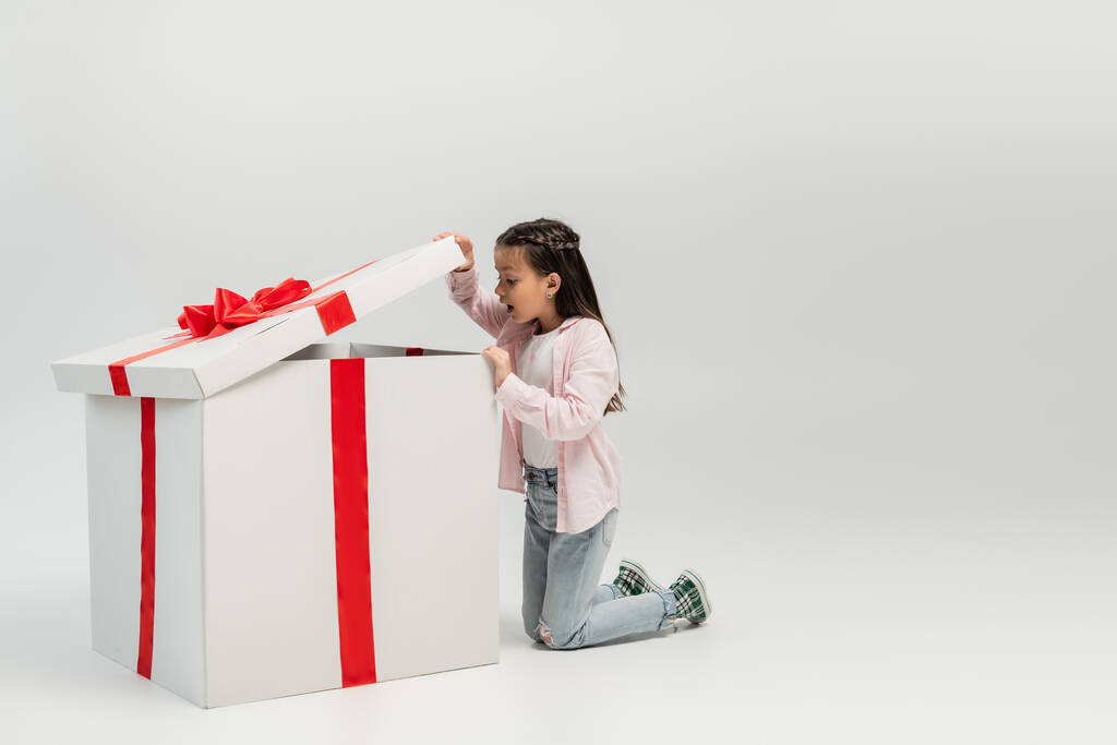 Vue latérale de l'enfant préadolescent choqué en vêtements décontractés ouvrant et regardant une grande boîte cadeau pendant la célébration de la journée internationale des enfants sur fond gris - Photo, image