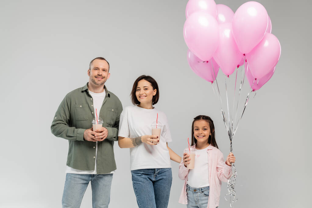 pais sorridentes e filha pré-adolescente segurando balões rosa e batidos enquanto olha para a câmera durante a celebração do dia internacional das crianças isolado em cinza  - Foto, Imagem