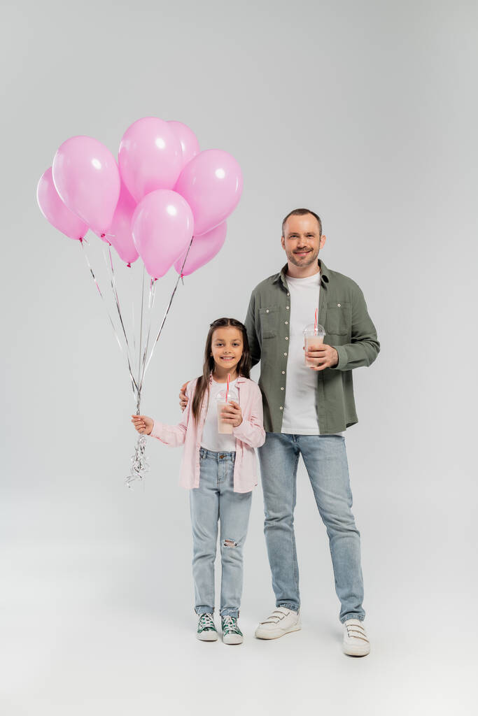 Χαμογελαστός πατέρας και προεφηβική κόρη σε casual ρούχα κρατώντας milkshakes και ροζ μπαλόνια, ενώ γιορτάζει την ημέρα προστασίας των παιδιών και στέκεται σε γκρι φόντο - Φωτογραφία, εικόνα