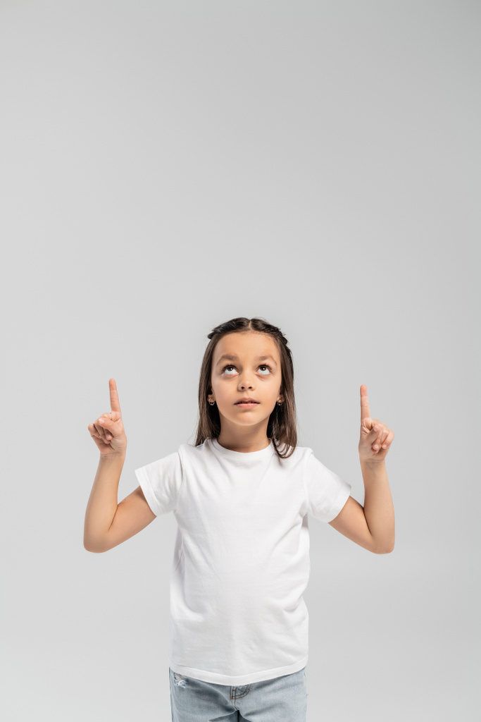 Menina pré-adolescente focada em camiseta branca e jeans apontando com os dedos e olhando para cima durante a celebração internacional do dia de proteção infantil isolada em cinza com espaço de cópia  - Foto, Imagem
