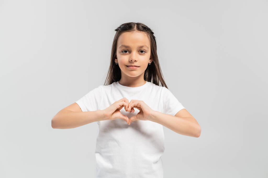 щаслива дівчина з довгим брюнетковим волоссям, що стоїть у білій футболці і показує жест серця з руками ізольовано на сірому фоні, день захисту дітей
  - Фото, зображення