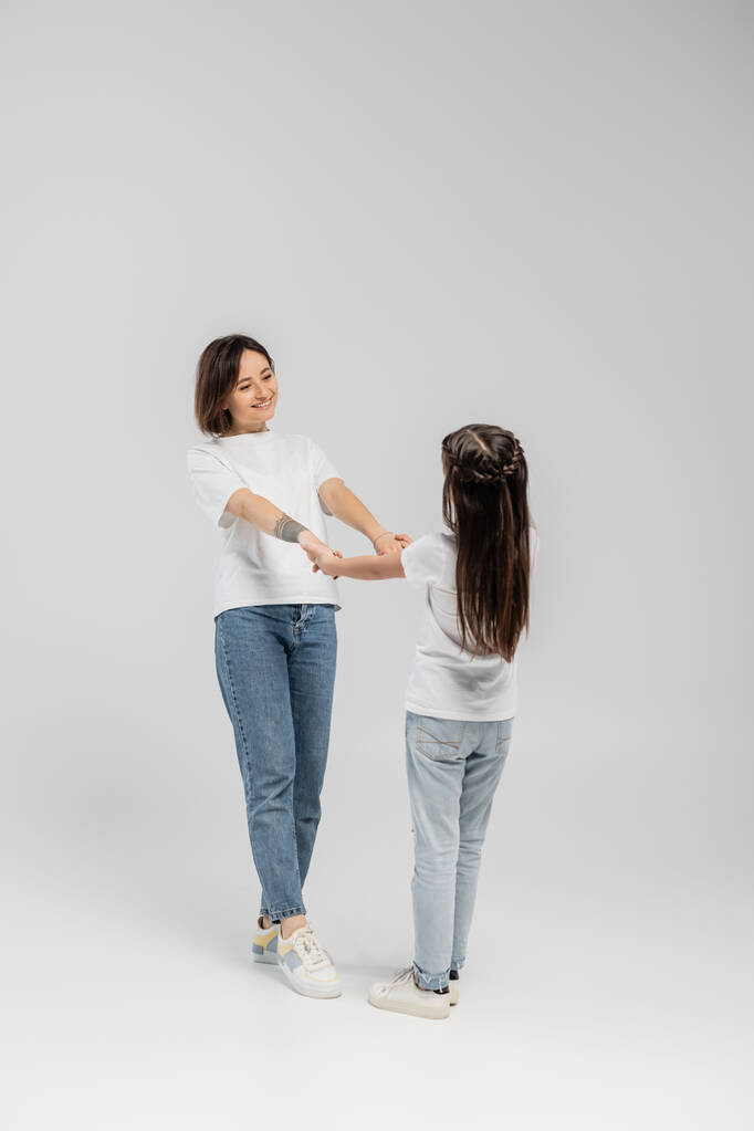 volledige lengte van de gelukkige moeder met tatoeage op de hand en kort haar hand in hand met tiener dochter terwijl ze samen staat in witte t-shirts en blauwe spijkerbroek op grijze achtergrond  - Foto, afbeelding