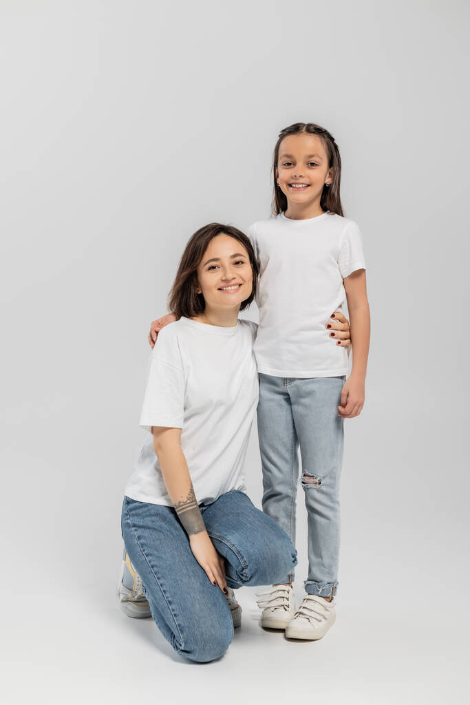 full length of happy mother with tattoo on hand and short hair hugging preteen daughter while posing together in white t-shirts and blue denim jeans on grey background  - Photo, Image