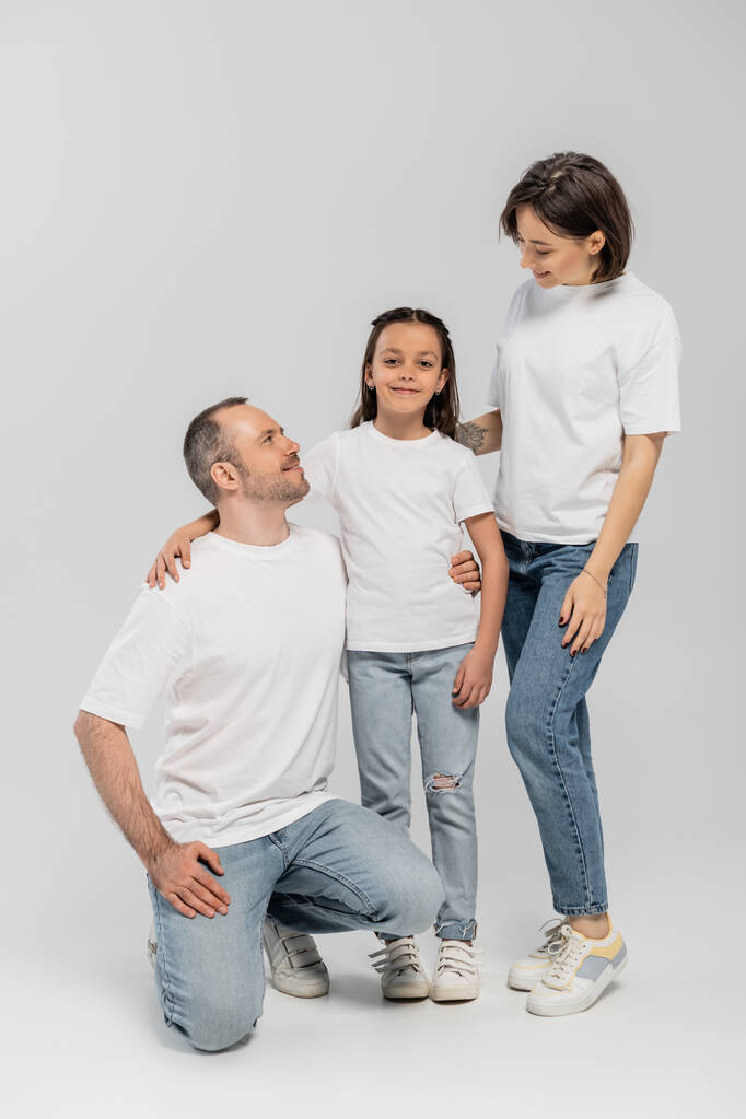 father and mother with short hair and tattoo on hand looking at cheerful preteen daughter while standing together in white t-shirts and blue denim jeans on grey background, child protection day  - Photo, Image