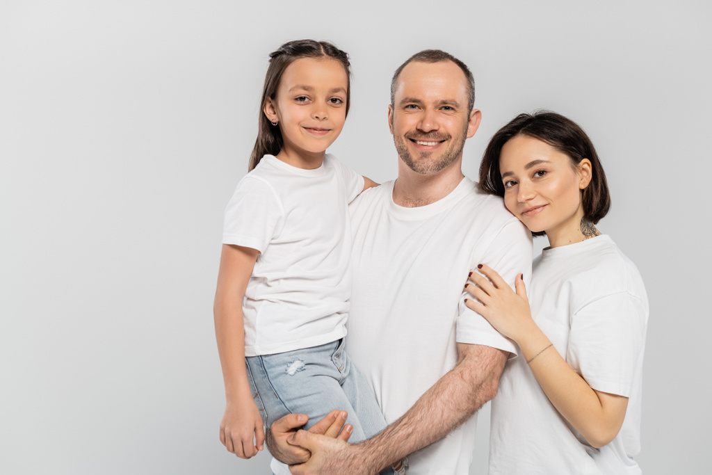 πορτρέτο της ευτυχισμένης οικογένειας σε λευκό t-shirts κοιτάζοντας κάμερα σε γκρι φόντο, Ημέρα προστασίας του παιδιού, χαρούμενο πατέρα ανύψωση preteen κόρη κοντά στη σύζυγο με κοντά μαλλιά και τατουάζ  - Φωτογραφία, εικόνα