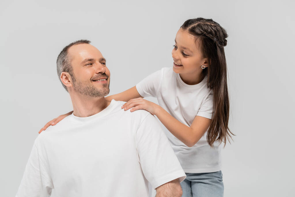 glückliches Mädchen mit langen brünetten Haaren, angelehnt an einen unrasierten und fröhlichen Vater mit Borsten, während sie in weißen T-Shirts posiert und sich auf grauem Hintergrund anschaut, Kinderschutztag  - Foto, Bild
