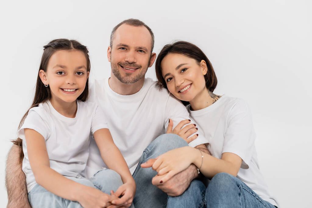 portrait of happy family in white t-shirts and blue denim jeans looking at camera and sitting together on grey background, International child protection day, parents and daughter  - Photo, Image