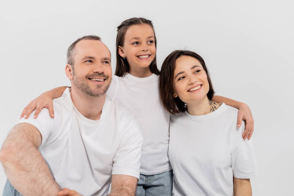 стильная семья в белых футболках смотрит в сторону, позируя вместе на сером фоне, Международный день защиты детей, дочь-подросток обнимает счастливых родителей  - Фото, изображение