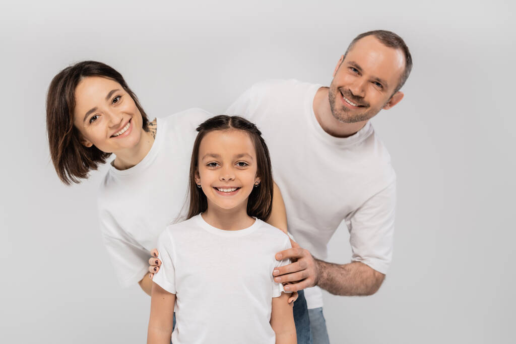 happy family in white t-shirts looking at camera and posing together on grey background, International child protection day, positive father mother and daughter  - Photo, Image