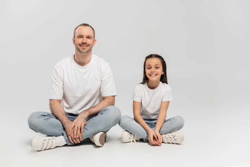 happy preteen girl with long brunette hair and cheerful dad with bristle sitting with crossed legs while posing in white t-shirts and blue denim jeans on grey background, Happy Father`s Day - Photo, Image