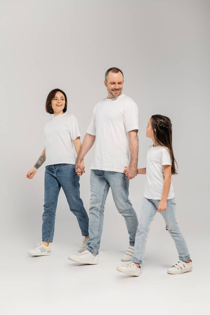 plná délka veselých rodičů a předpubertální dcery v bílých tričkách a modrých džínách džínách držící se za ruce a kráčející společně na šedém pozadí, Mezinárodní den ochrany dětí v červnu  - Fotografie, Obrázek
