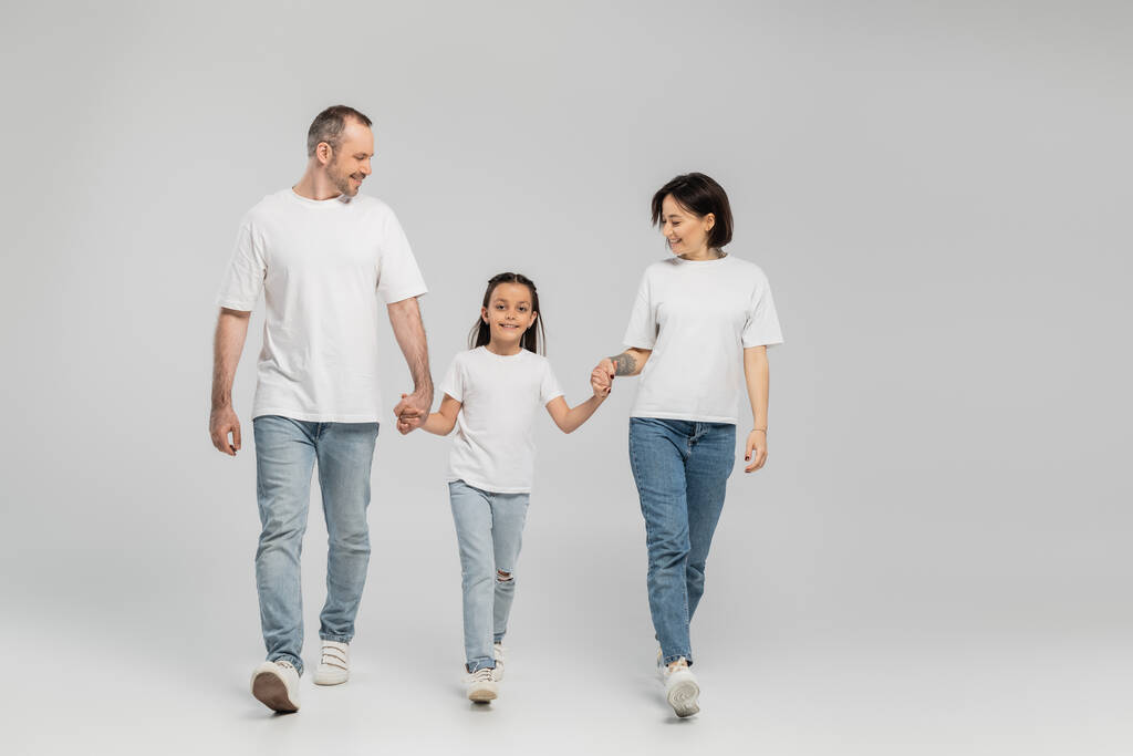 полная длина счастливых родителей и ребенка в белых футболках и джинсах в синих джинсах, держащихся за руки и идущих вместе на сером фоне, Международный день защиты детей в июне  - Фото, изображение