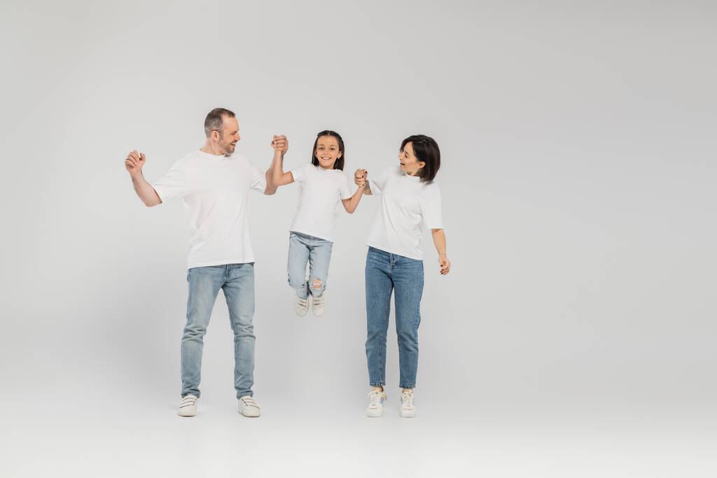 onnellisten vanhempien täysi pituus valkoisissa t-paidoissa ja sinisissä farkuissa, jotka pitelevät toisiaan kädestä ja nostavat esiteini-ikäistä tytärtä seistessään yhdessä harmaalla pohjalla, kansainvälinen lastensuojelupäivä kesäkuussa  - Valokuva, kuva