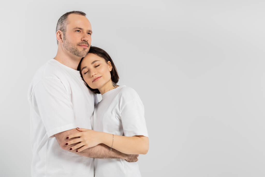 tetovaná žena s krátkými brunetovými vlasy a zavřenýma očima, opírající se o hruď manžela se štětinami, zatímco stojí spolu v bílých tričkách izolovaných na šedém pozadí, šťastný pár  - Fotografie, Obrázek