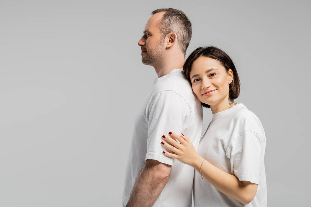 татуированная женщина с короткими волосами брюнетки опираясь на спину мужа, стоя вместе в белых футболках и глядя на камеру изолированы на сером фоне, счастливая пара  - Фото, изображение