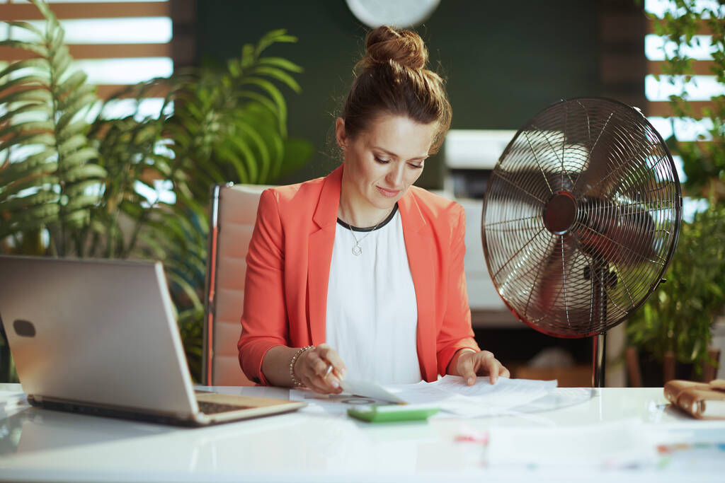 Kestävä työpaikka. moderni 40-vuotias kirjanpitäjä nainen modernissa vihreässä toimistossa punaisessa takissa, jossa on sähköinen tuuletin ja kannettava tietokone. - Valokuva, kuva