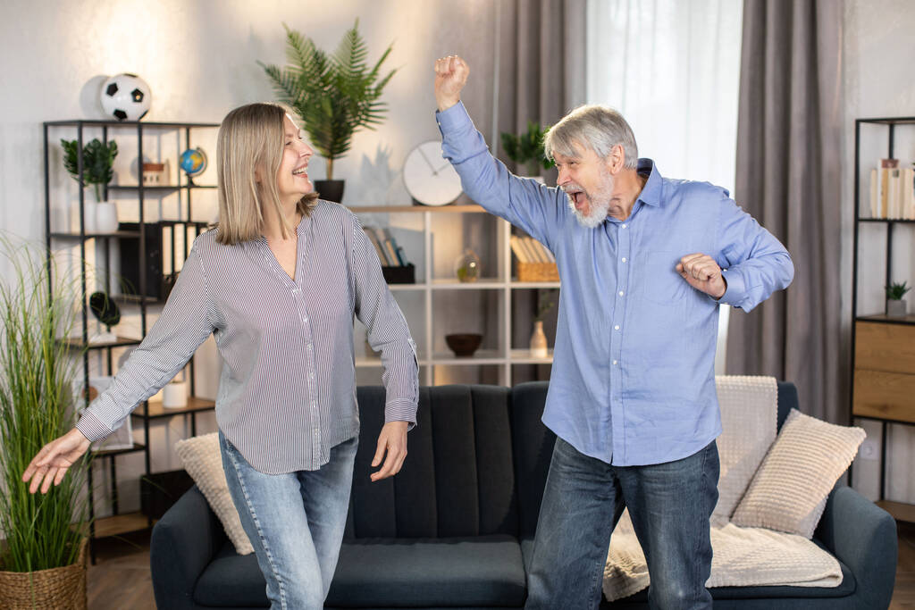 Irres Rentnerpaar in Alltagskleidung tanzt munter zusammen im modernen Wohnzimmer. Unbekümmerte Eheleute, die sich in der Freizeit ausruhen und das Leben in jedem Alter genießen. - Foto, Bild