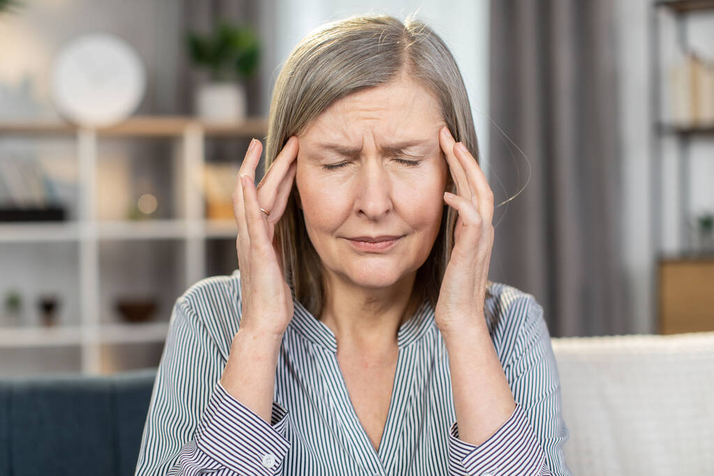 Frustrierte Seniorin leidet unter starker plötzlicher Migräne und Schwindel, während sie auf der Couch sitzt. Kranke alte Frau in gestreiftem Hemd massiert Schläfen mit den Fingern im Haus. Gesundheitskonzept. - Foto, Bild