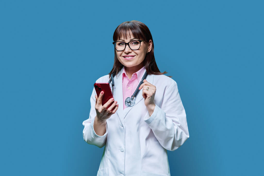 Starší doktorka v bílém laboratorním plášti s telefonem, na modrém pozadí studia. Medicína, on-line internetová služba, povolání, zdravotní péče, zdravotní péče, profesionální mobilní aplikace, koncepce léčby - Fotografie, Obrázek