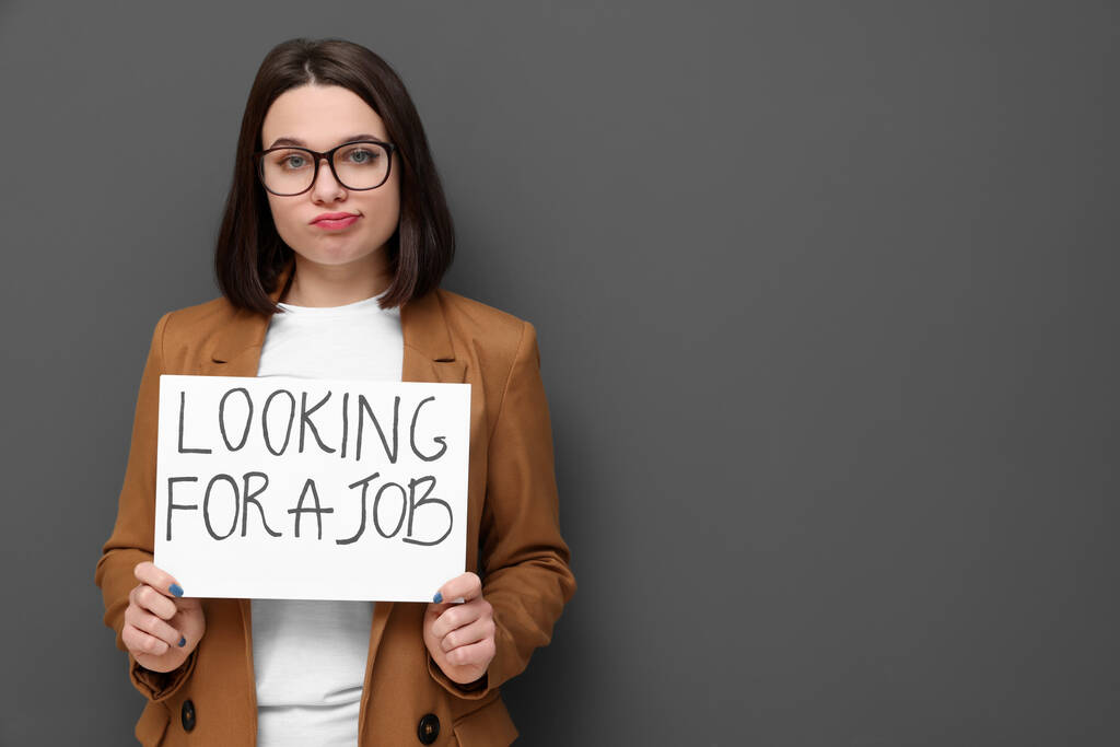 若い失業女性は灰色の背景に仕事を探してフレーズで看板を保持動揺します.テキストのスペース - 写真・画像