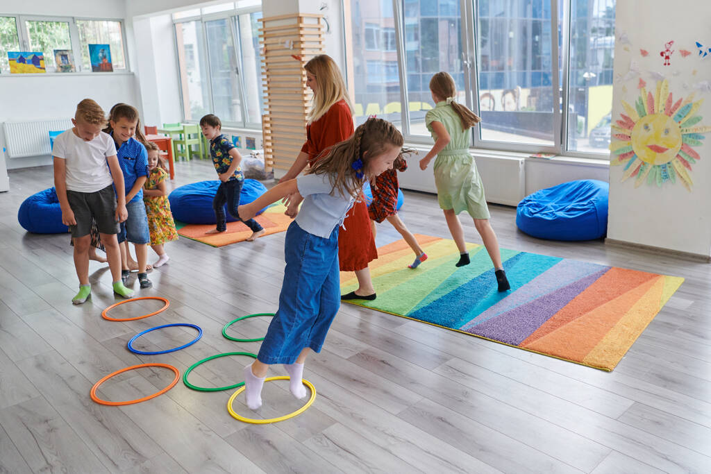 Malé děti z mateřské školy s učitelkou na podlaze ve třídě, cvičí. Skákání přes kruhy hula obruče na podlaze - Fotografie, Obrázek