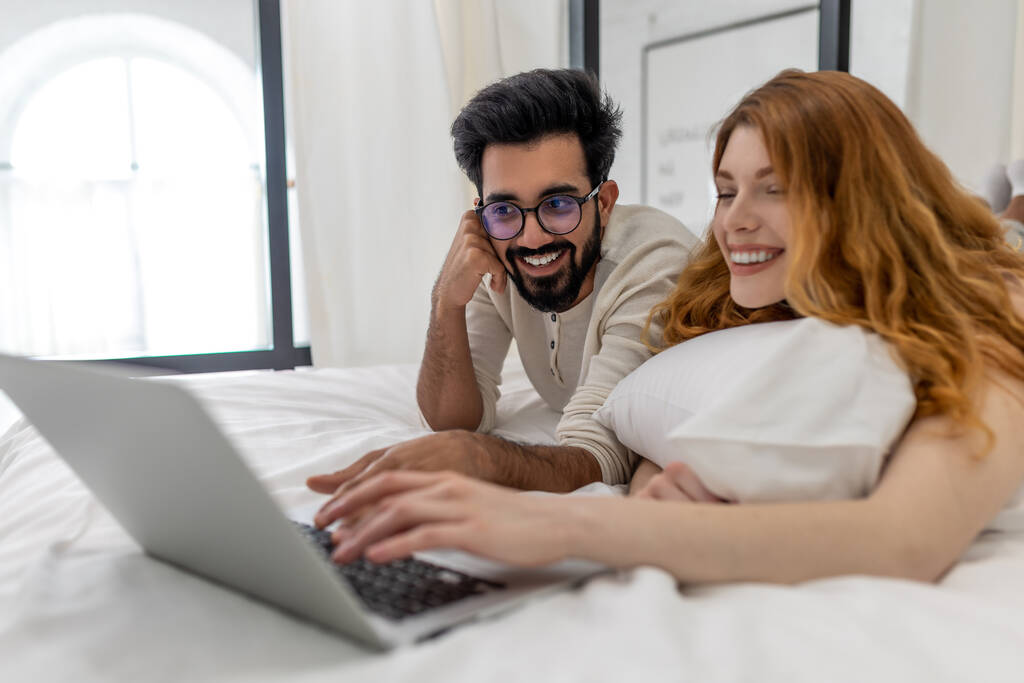Szczęśliwa radosna żona i mąż za pomocą laptopa w sypialni, przeglądanie Internetu razem lub oglądanie filmu. - Zdjęcie, obraz