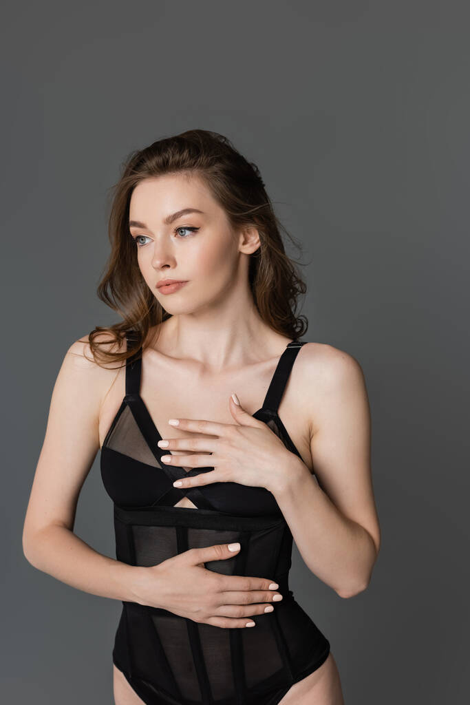 Portrait of sexy and brunette woman with natural makeup posing in black and trendy, form-fitting bodysuit and touching chest while looking away and standing isolated on grey   - Photo, Image