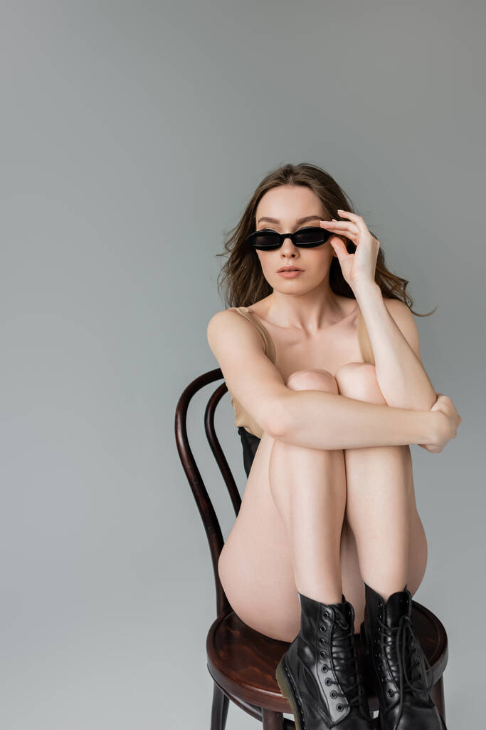 Giovane donna alla moda con acconciatura indossando body e stivali neri e toccando occhiali da sole alla moda mentre seduta su sedia in legno isolata su grigio   - Foto, immagini