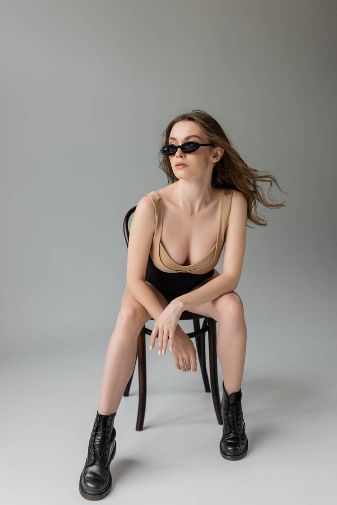 Full length of trendy νεαρή μελαχρινή μοντέλο σε γυαλιά ηλίου, μπεζ bodysuit, μαύρο κορσέ και μπότες που ποζάρουν ενώ κάθονται σε ξύλινη καρέκλα σε γκρι φόντο  - Φωτογραφία, εικόνα