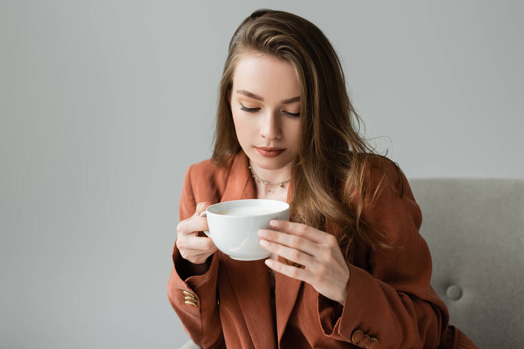 Trendige brünette junge Frau in Halskette und Terrakottamaske, die eine Tasse Cappuccino hält, während sie auf einem modernen Sessel sitzt, isoliert auf grau mit Kopierraum - Foto, Bild
