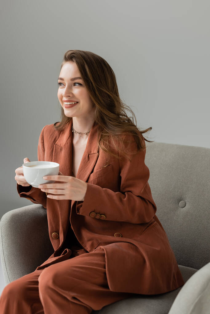 Fröhliche junge und modische Frau in Terrakotta-Anzug und Halskette hält eine Tasse Cappuccino in der Hand und schaut weg, während sie es sich auf einem modernen Sessel auf grau isoliert bequem macht   - Foto, Bild