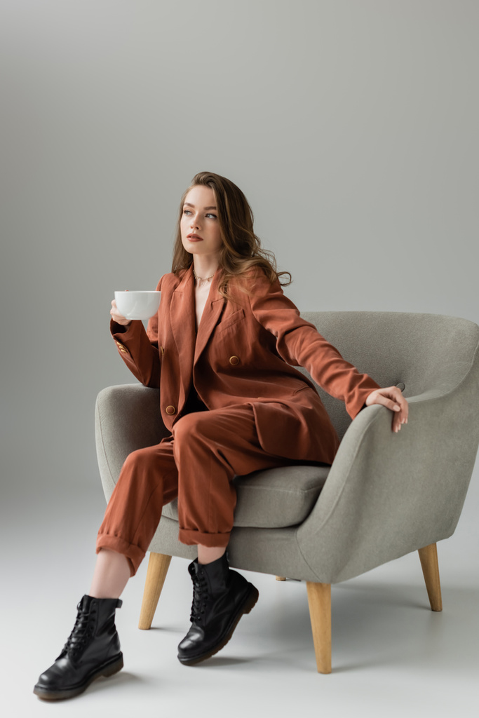 brunette young woman with long hair and necklace wearing terracotta and trendy suit with blazer and pants and holding cup of coffee while sitting in comfortable armchair on grey background in studio  - Photo, Image