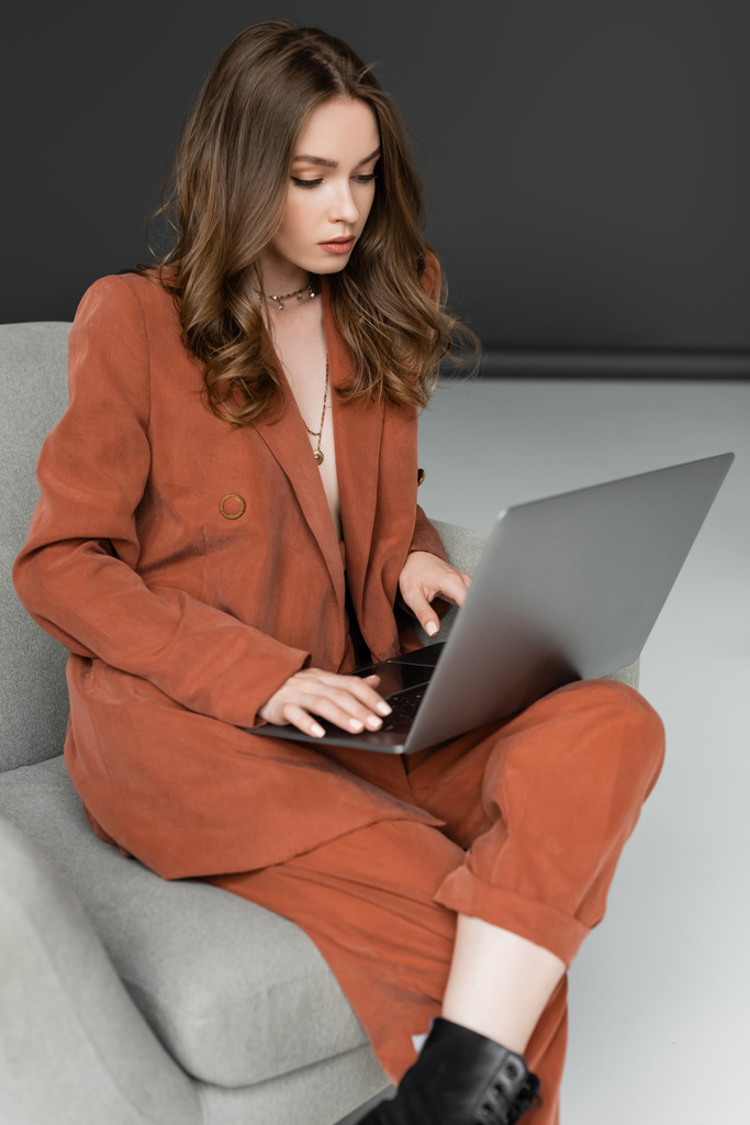 брюнетка молодая женщина с длинными волосами и ожерелье носить модный костюм с блейзером и брюки с помощью ноутбука, сидя в удобном кресле на сером фоне, фрилансер, удаленная работа  - Фото, изображение