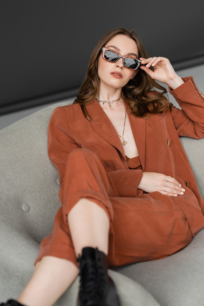 stylish young woman with long hair wearing terracotta suit with blazer and pants and posing in trendy sunglasses while sitting in blurred boots on armchair on grey background, fashionable model  - Photo, Image