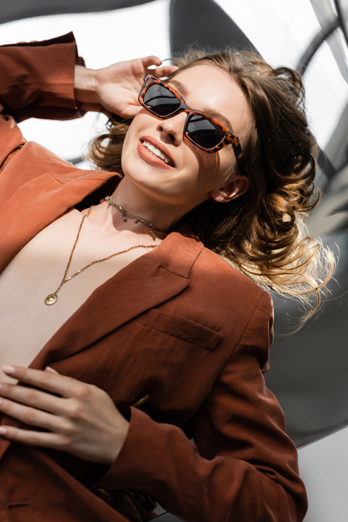 top view of positive woman with long hair wearing terracotta and trendy suit with blazer and golden necklace and posing in sunglasses on abstract grey background, beautiful model, sunlight  - Photo, Image