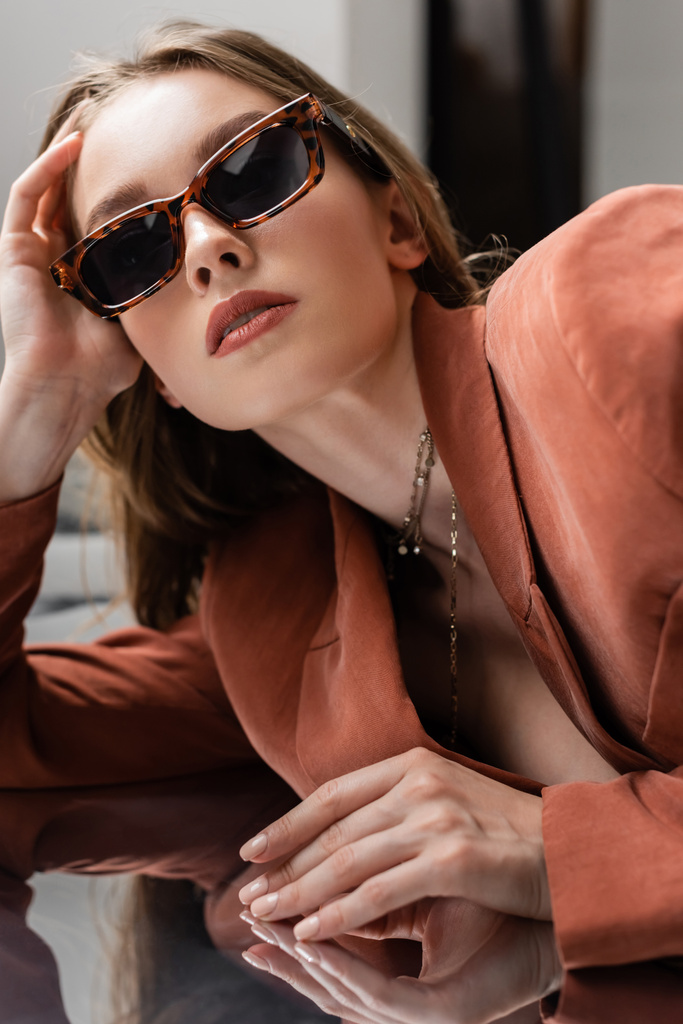 junge Frau in Terrakotta und trendigem Anzug mit Blazer und goldener Halskette und posiert in modischer Sonnenbrille auf grau verspiegeltem Hintergrund, schönes Modell, Spiegelung, Blick in die Kamera - Foto, Bild