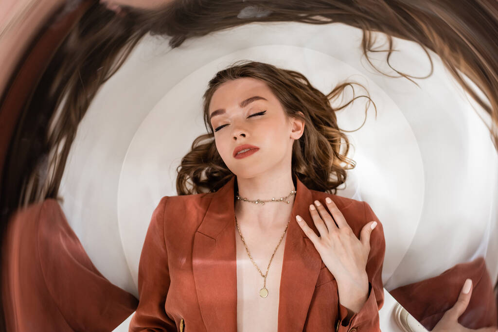 circular shot of young woman with brunette hair and closed eyes wearing blazer and golden necklace while lying on grey background, blurred, wide-angle, distorted view, top view, creative perspective  - Photo, Image