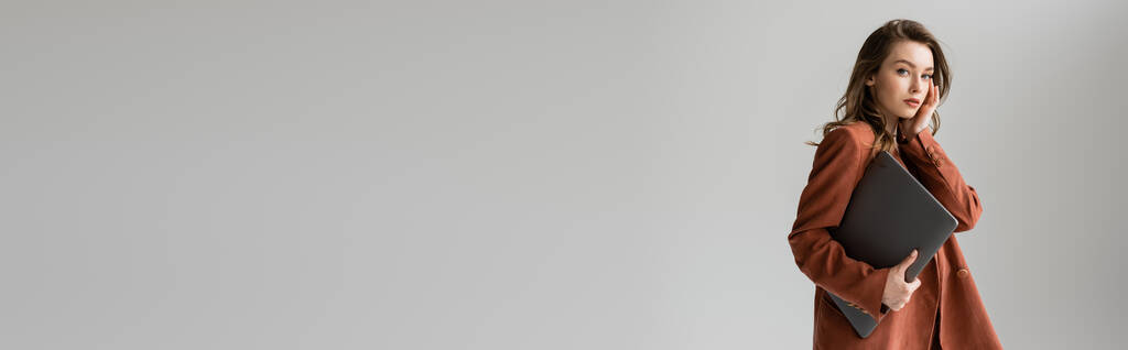 giovane donna con capelli castani e collana indossando abito alla moda in terracotta con blazer, guardando la fotocamera e tenendo il computer portatile mentre in piedi su sfondo grigio, freelance, lavoro a distanza, banner - Foto, immagini