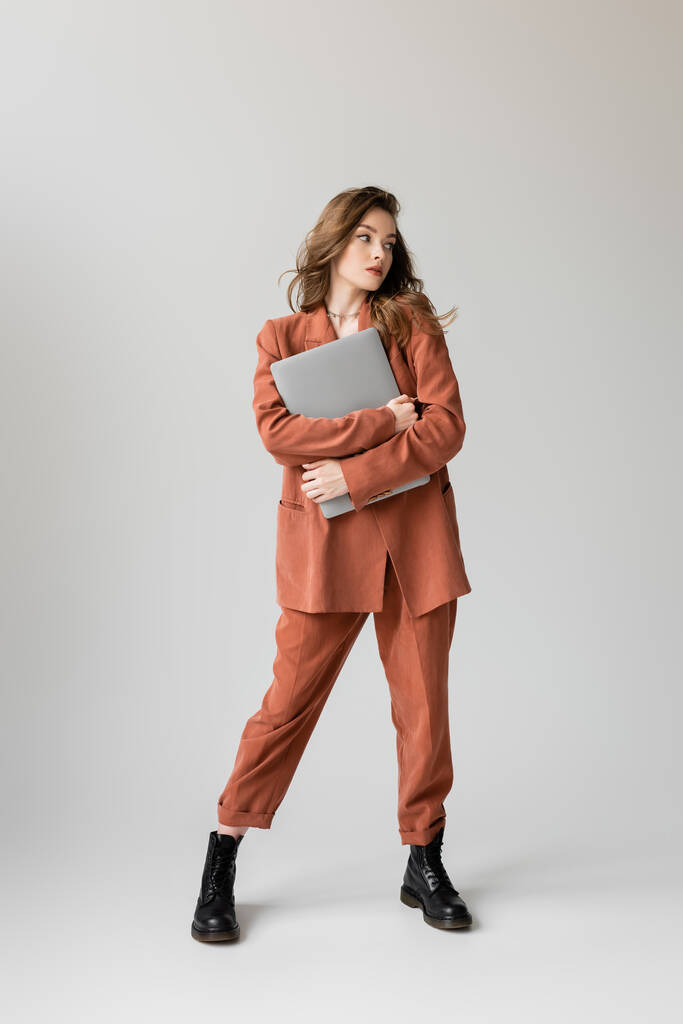 full length of young woman with brunette hair and necklace wearing terracotta and trendy suit with blazer and pants, holding laptop while standing on grey background, freelancer, remote work  - Photo, Image
