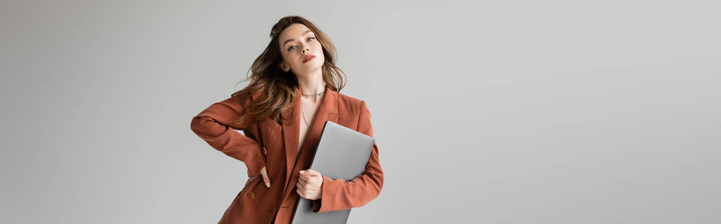 modny freelancer z brunetką włosy w brązowym i modnym garniturze z marynarką, trzymając laptopa stojąc z ręką na biodrze na szarym tle, patrząc aparat fotograficzny, młoda kobieta, praca zdalna, baner  - Zdjęcie, obraz