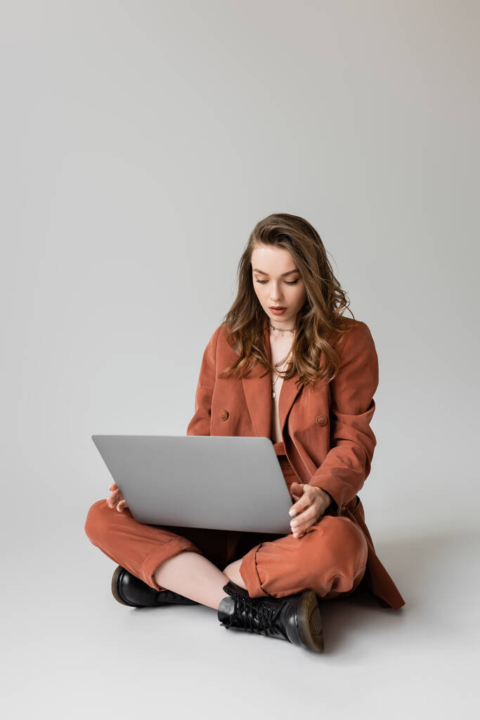 brunetka mladá žena ve zlatém náhrdelníku sedí se zkříženýma nohama v terakotě a módní oblek, pomocí notebooku při práci na dálku na šedém pozadí, na volné noze, digitální nomád  - Fotografie, Obrázek
